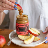 Person dipping an apple slice into a jar of Butter Bike peanut butter on a plate with more apple slices.